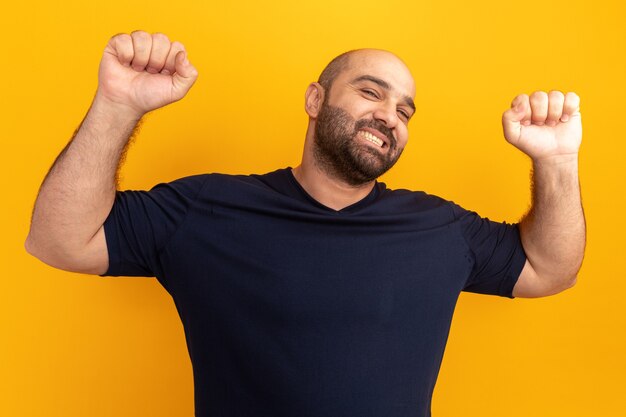 Счастливый и веселый бородатый мужчина в темно-синей футболке поднимает кулаки, как победитель, стоящий над оранжевой стеной
