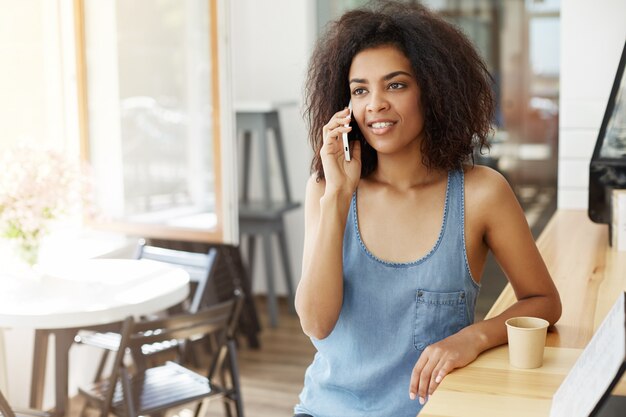 カフェに座って電話で話して笑って幸せな陽気な美しいアフリカ女性。