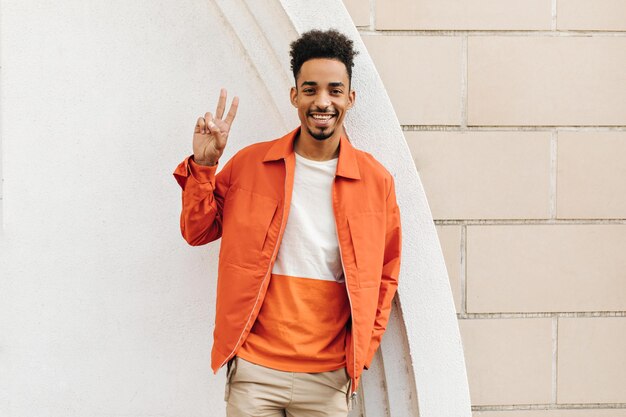 Happy cheerful brunet darkskinned man in orange jacket and beige shorts smiles sincerely and shows peace sign outside