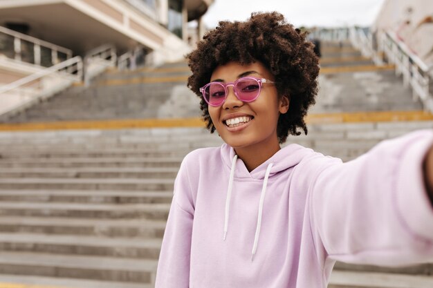 紫色の特大のパーカーとピンクのサングラスで幸せな陽気な巻き毛の女性は笑顔で階段の近くの外で自分撮りを取ります