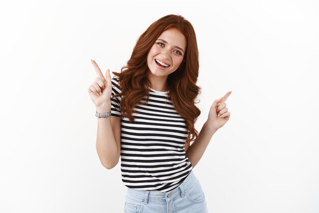 Happy cheerful ginger girl in striped tshirt dancing pointing upper left and right corners indicate promo smiling joyfully introduce advertisement recommend click offer white background