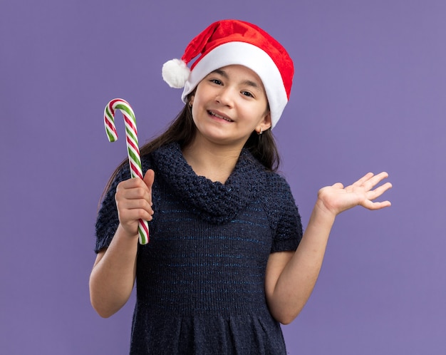 Happy and cheerful  little girl in knit dress wearing santa hat holding candy cane  with smile on face  standing over purple wall