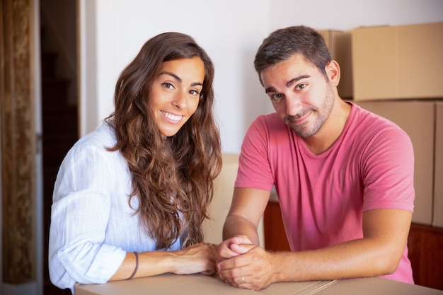 Happy cheerful man and woman enjoying moving into new house, standing indoors, leaning on carton box, 