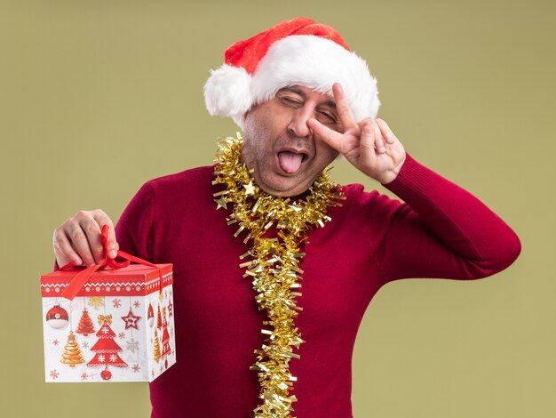 Счастливый и веселый мужчина среднего возраста в рождественской шляпе санта-клауса с мишурой на шее, держащий рождественский подарок, глядя в камеру, высунув язык, показывая v-знак, стоящий на зеленом фоне