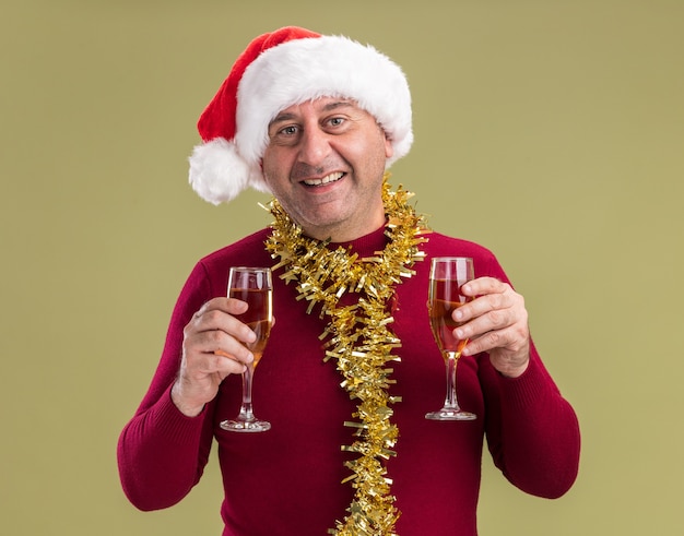 緑の背景の上に立って笑顔のカメラを見てシャンパンのグラスを持って首の周りに見掛け倒しのクリスマスサンタ帽子をかぶって幸せで陽気な中年男性