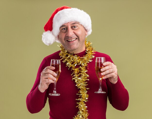 녹색 배경 위에 서 웃는 카메라를보고 샴페인 잔을 들고 목 주위에 반짝이와 크리스마스 산타 모자를 쓰고 행복하고 쾌활한 중년 남자