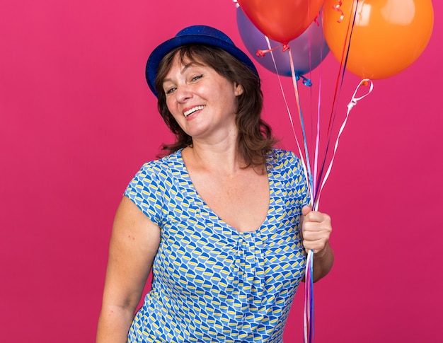 Счастливая и веселая женщина среднего возраста в партийной шляпе, держащая кучу разноцветных шаров, широко улыбаясь, празднует день рождения, стоя над розовой стеной