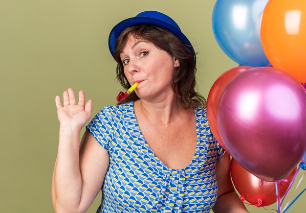 Happy and cheerful middle age woman in party hat with bunch of colorful balloons blowing a whistle