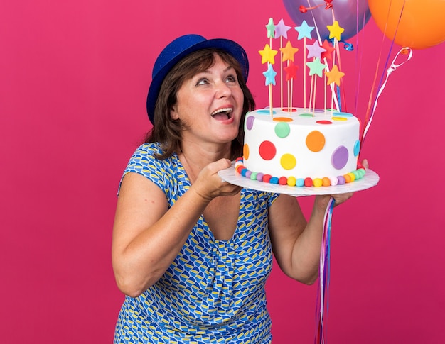 Happy and cheerful middle age woman in party hat with colorful balloons holding birthday cake looking up smiling broadly celebrating birthday party standing over pink wall