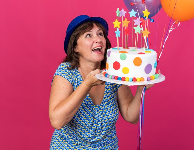 생일 케이크를 들고 다채로운 풍선과 함께 파티 모자에 행복하고 쾌활한 중년 여자는 분홍색 벽 위에 서 생일 파티를 광범위하게 축하 미소를 찾고