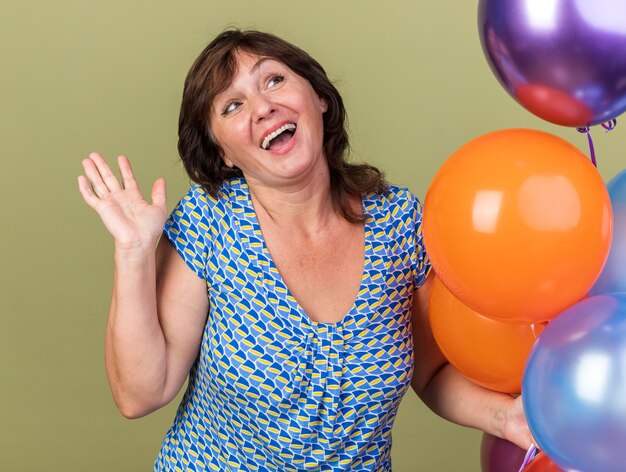 Happy and cheerful middle age woman with bunch of colorful balloons looking up smiling broadly with arm raised