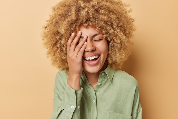 Happy cheerful woman makes face palm laughs joyfully expresses sincere feelings and positive emotions dressed in casual linen shirt isolated over brown background Peope and happiness concept
