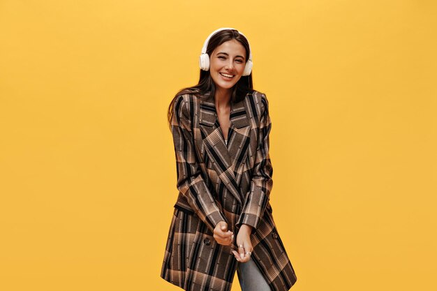 Happy cheerful woman in oversized brown long jacket laughs on isolated Beautiful brunette girl smiles and listens to music in headphones on yellow background