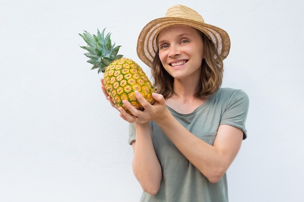 Happy cheerful woman in summer hat showing whole pineapple fruit