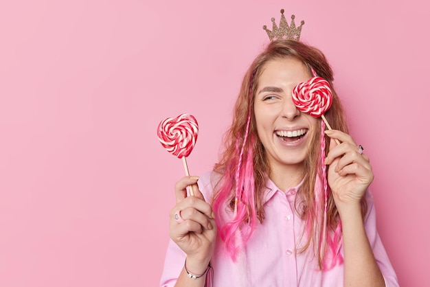 Happy cheerful woman with long hair covers eyes with sweet candies on sticks has upbeat mood smiles broadly wears little crown and shirt isolated over pink background blank copy space on left