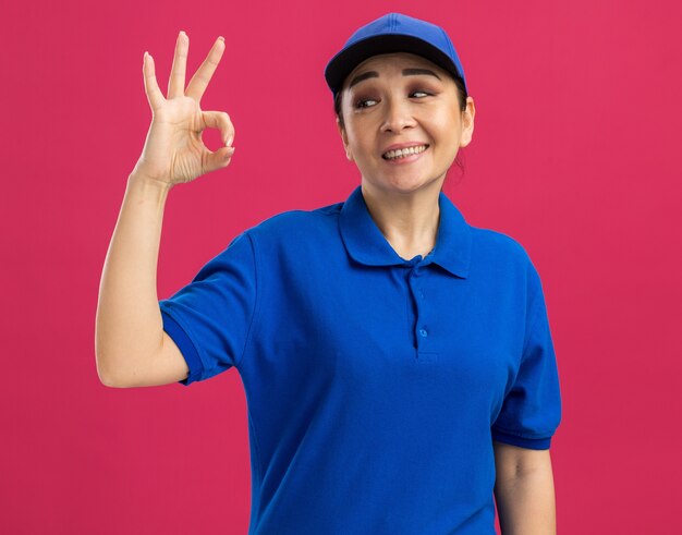 Счастливая и веселая молодая женщина-доставщик в синей форме и кепке улыбается, делая знак ОК