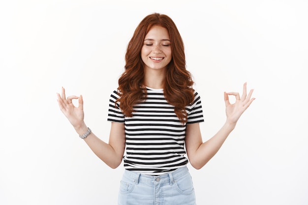 Happy cheerful young ginger girl with freckles, long red hairstyle, close eyes and smiling dreamy, feeling relieve and happiness as meditating, breathing exercises, show zen mudra sign, calm down