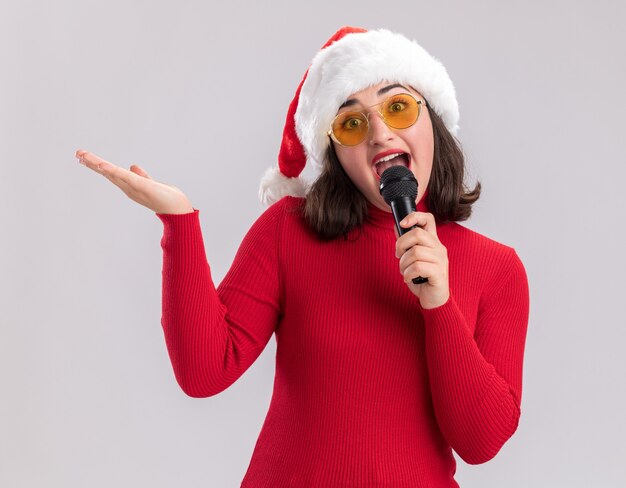 Happy and cheerful young girl in red sweater and santa hat wearing glasses holding microphone looking at camera smiling with arm raised standing over white background