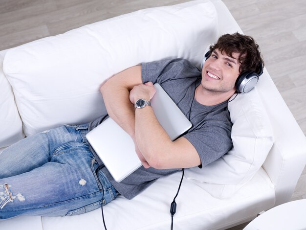 抱擁のラップトップとヘッドフォンのソファーに横になっている幸せな明朗快活な青年