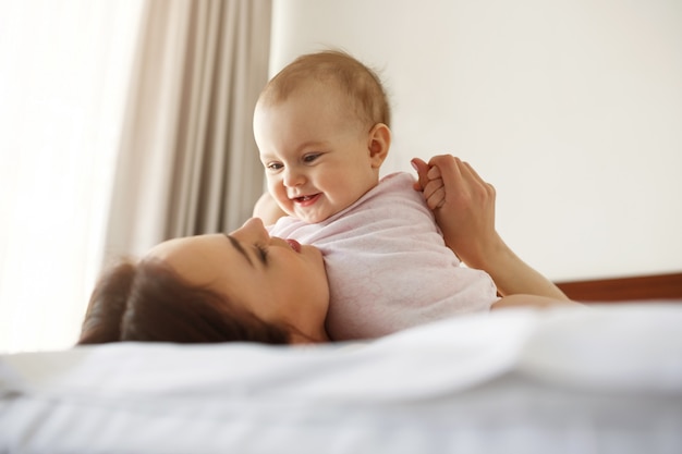 Happy cheerful young mom smiling playing with her little baby daughter lying in bed at home.