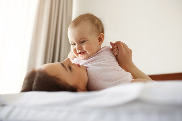 Счастливая жизнерадостная молодая мама усмехаясь играющ при ее маленькая дочь младенца лежа в кровати дома.