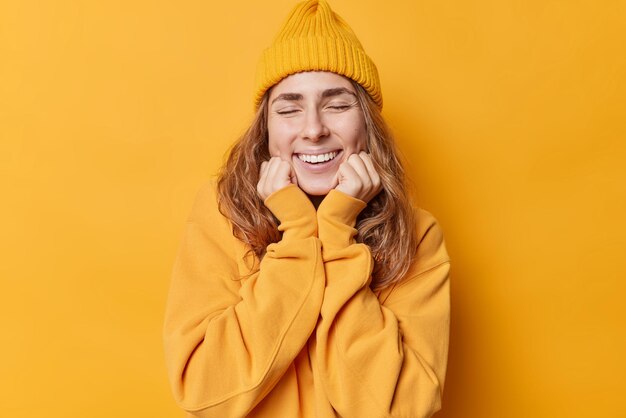 Happy cheerful young woman smiles gently daydreams about something keeps hands under chin keeps eyes closed dressed in casual hoodie and hat isolated over yellow background Pleasant feelings