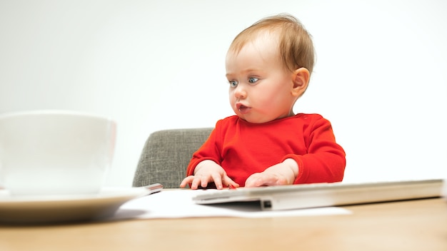 Neonata felice del bambino che si siede con la tastiera del calcolatore o del laptop moderno isolata su uno studio bianco.