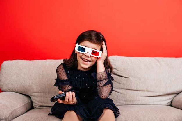 Free photo happy child enjoying tv on sofa indoor shot of little girl in 3d glasses sitting on red background