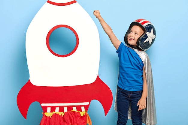 幸せな子供は紙のカートンのおもちゃのロケットの近くで腕を上げ、宇宙に飛びたい