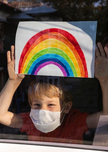 Happy child with medical mask holding rainbow sign through the window at home