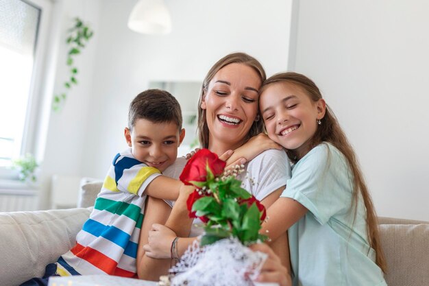 Happy children giving gift an flowers to mother Happy Mothers Day Children boy and girl congratulate smiling mother give her flowers bouquet of roses and a gift box during holiday celebration