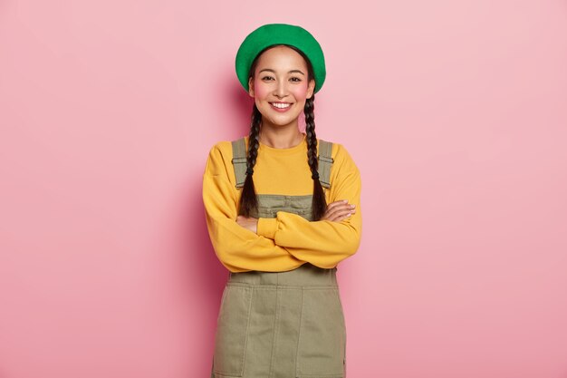 Happy Chinese millennial girl keeps arms folded, smiles pleasantly at camera, enjoys pleasant talk with boyfriend, wears green beret, yellow sweatshirt