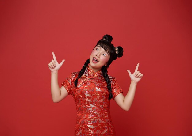 Happy Chinese new year Asian child girl wearing traditional cheongsam qipao dress and hands pointing finger up isolated on red copy space background