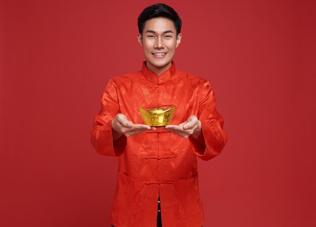 Free photo happy chinese new year. asian man wearing in traditional costume holding gold ingot isolated on red background.