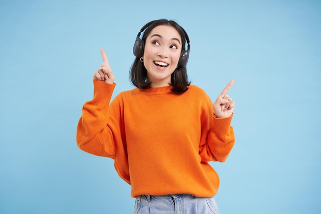 Free photo happy chinese woman in headphones listens music enjoys favourite song in her playlist stands over bl