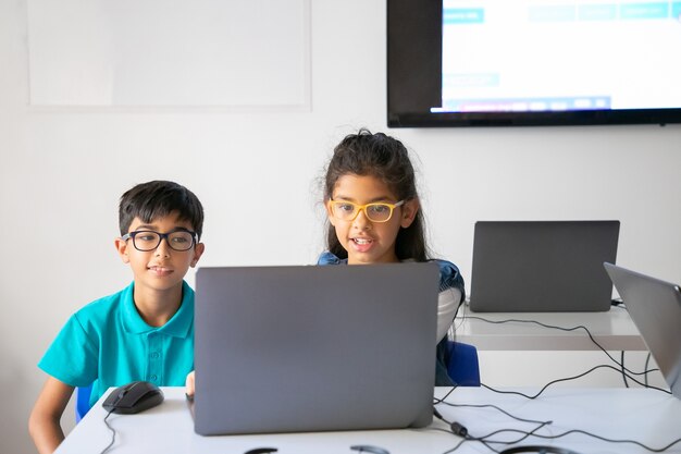 Happy classmates in glasses sitting at table together and using laptop in classroom