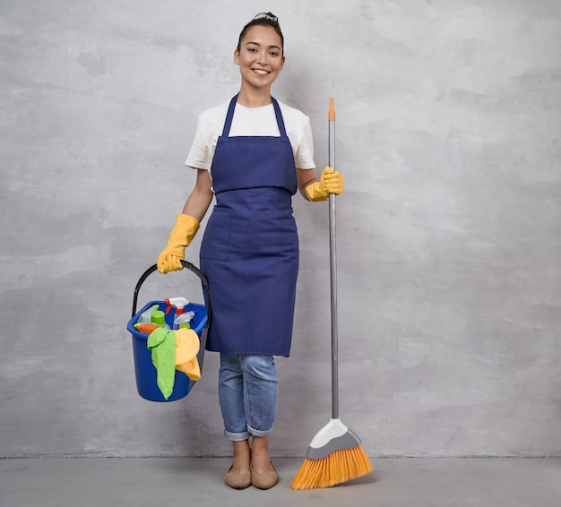 Счастливая уборщица в униформе и желтых резиновых перчатках держит метлу и пластиковое ведро с Premium Фотографии