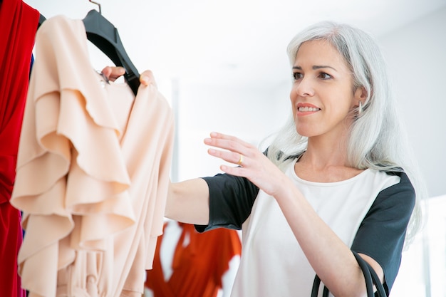 幸せな洋服店のお客様は、試着のためにラックからドレスでハンガーを取ります。ファッション店で洋服を選ぶ女性。消費主義または小売の概念