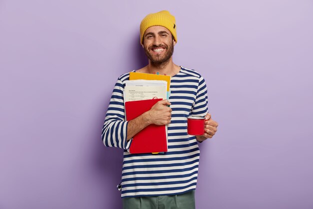 Happy college student carries papers and notepad, prepares for exams, drinks hot beverage from cup