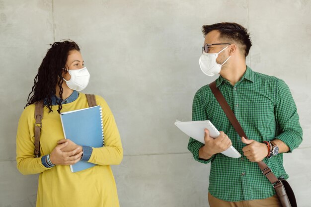 Happy college students wearing protective face masks while standing by the wall and talking.