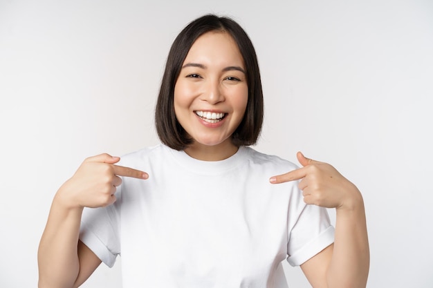 Felice e sicura di sé studentessa asiatica sorridente e indicando se stessa autopromozione che mostra il logo sulla maglietta in piedi su sfondo bianco