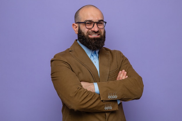 Happy and confident bearded man in brown suit wearing glasses looking at camera smiling cheerfully with arms crossed standing over purple background