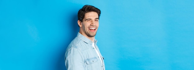 Happy and confident man turn head at camera winking and smiling standing over blue background