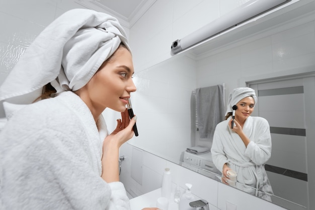 Счастливая уверенная молодая дама с полотенцем на голове делает макияж