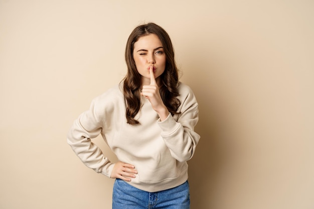 Happy coquettish girl say shh, shushing, keep secret, be quiet gesture, smiling at camera, standing over beige background