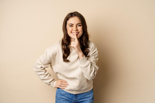 Happy coquettish girl say shh, shushing, keep secret, be quiet gesture, smiling at camera, standing over beige background