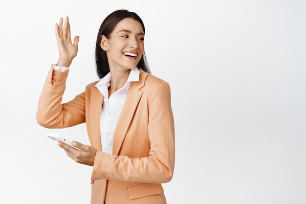 Happy corporate woman using smartphone turn behind to wave at friend saying hello to coworker while passing by with mobile phone white background