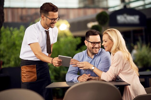 Happy couple choosing order while waiter is showing the menu on digital tablet in a restaurant