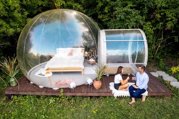 Free photo a happy couple drinking wine near transparent bubble tent at glamping nature around