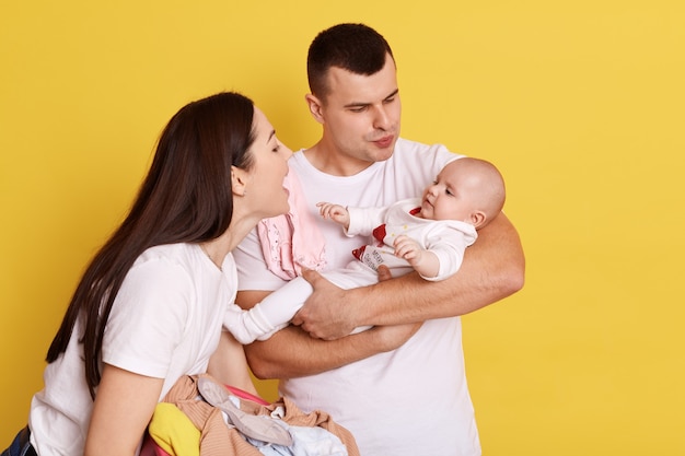 黄色の背景の上に生まれたばかりの子供を抱きしめて見ている幸せなカップル、愛と笑顔で小さな娘と話している、白いTシャツを着ている両親、屋内で幸せな家族。